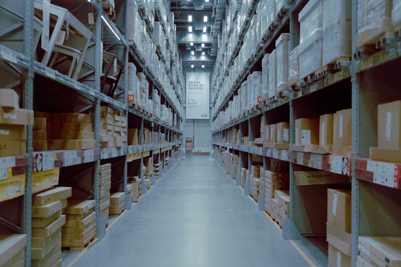 Long stockroom aisle lined with shelving bays holding boxed inventory on both sides.